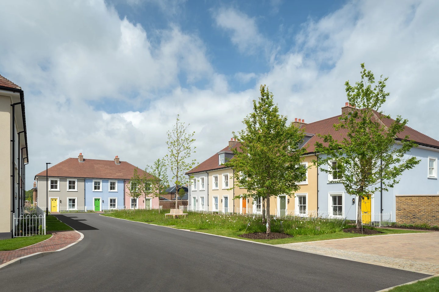 Row of pastel coloured houses with white windows and coloured front doors along a curved residential street with trees and grass