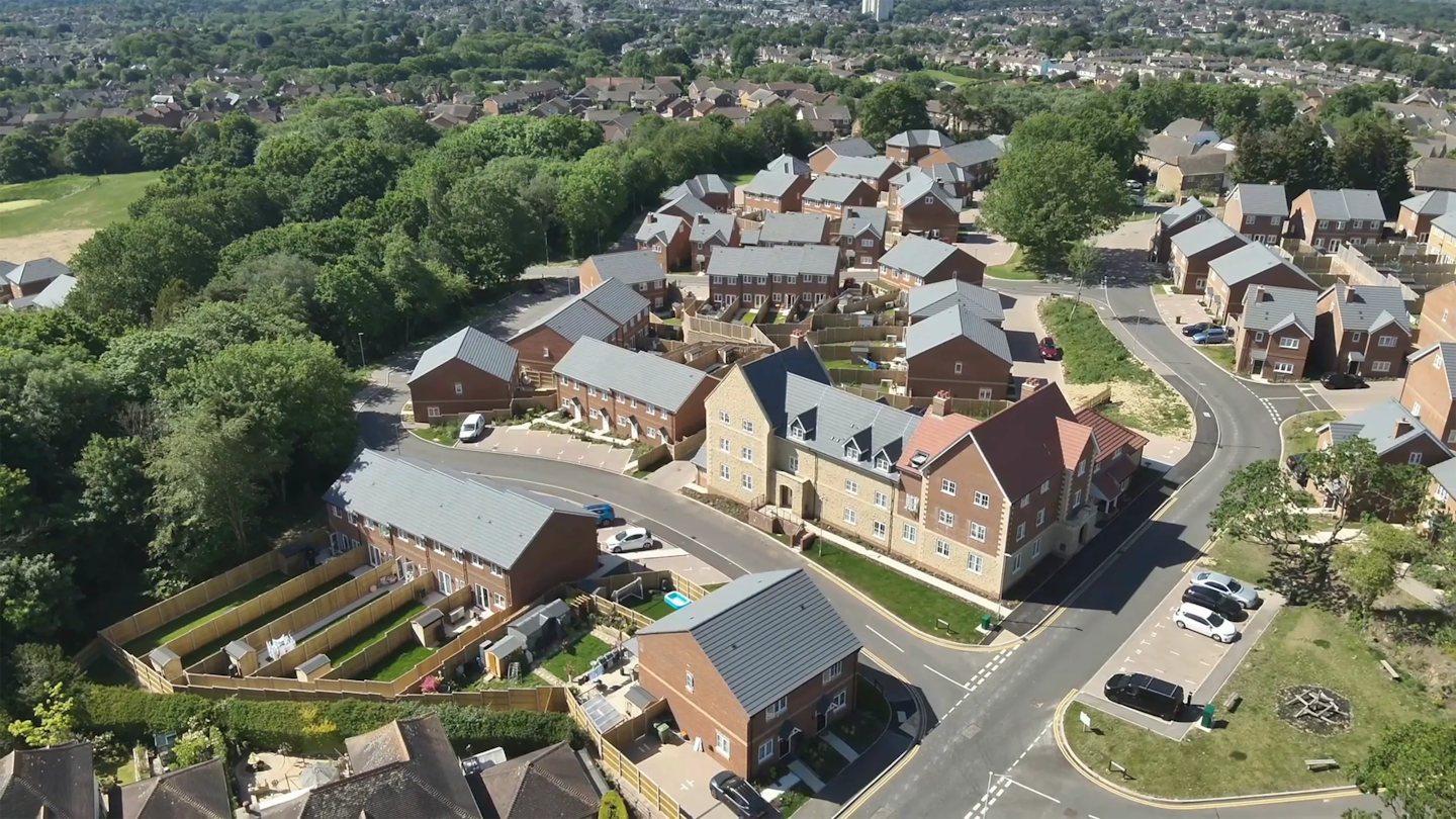 Aerial view of a residential area showing a mix of houses and apartments centred around a main road junction, with nearby greenery.