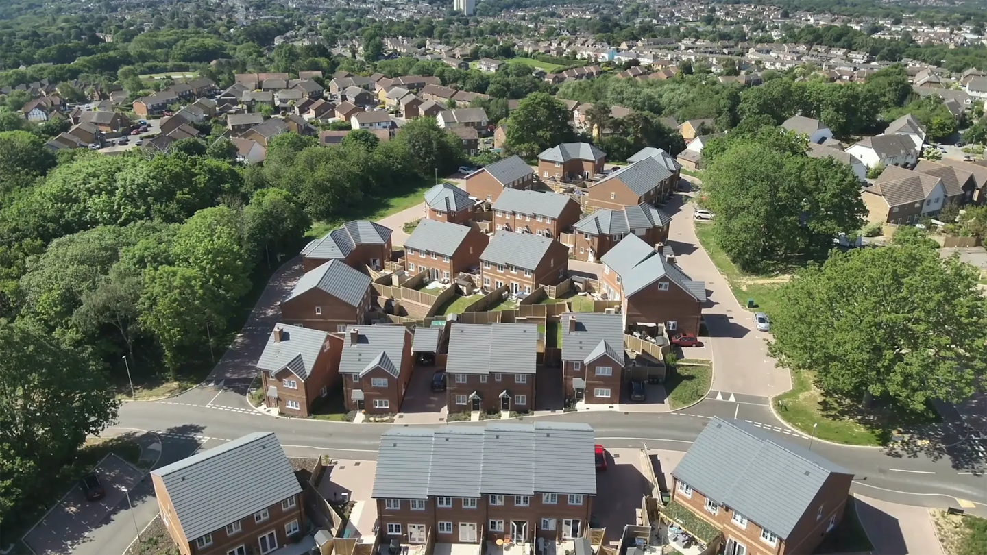 Wider aerial view of a completed development with mixed housing types, surrounding woodland and suburban neighbourhood beyond.