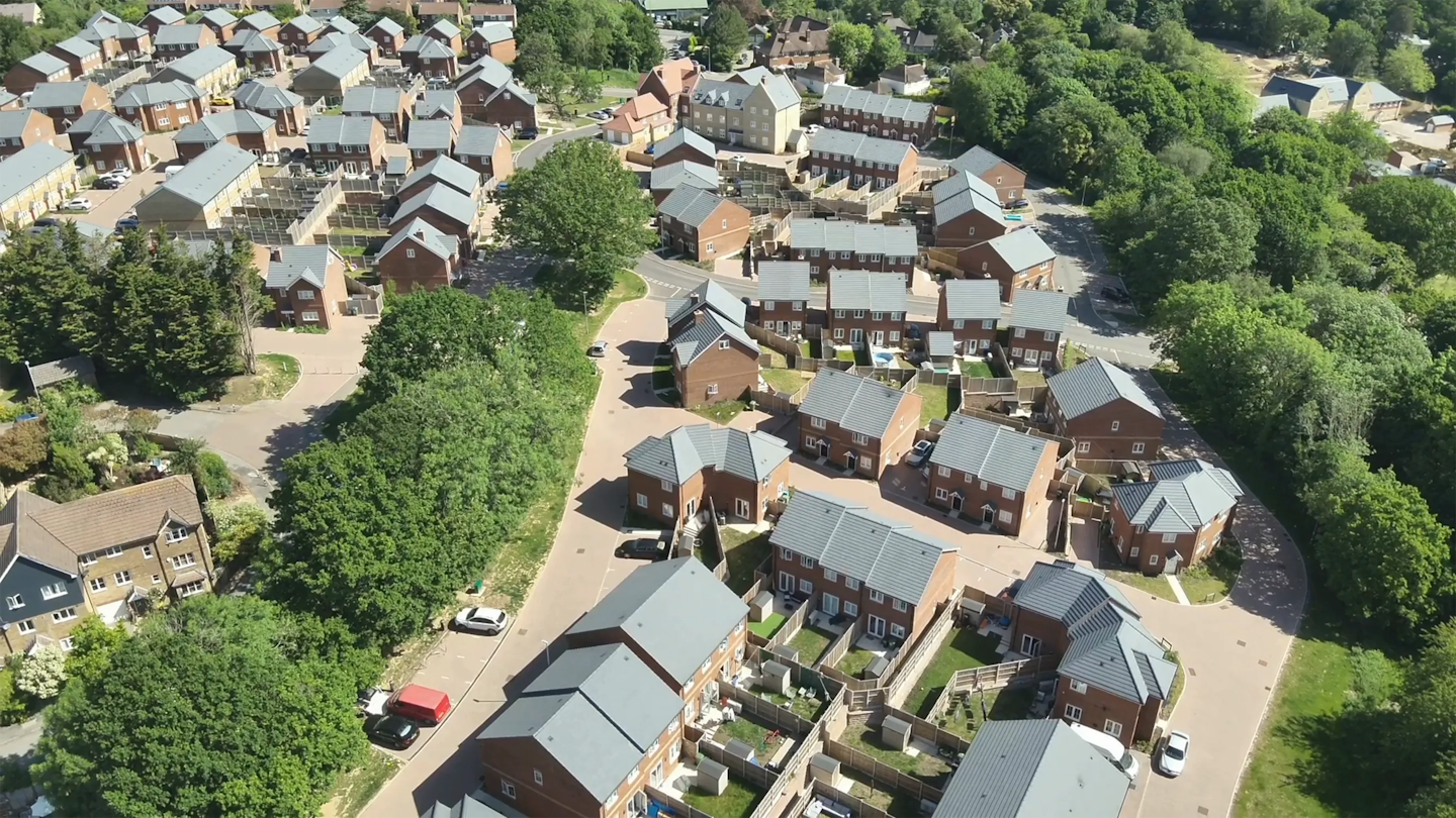 Aerial view showing a housing development next to mature trees and open green space, with homes arranged around gently curving roads.