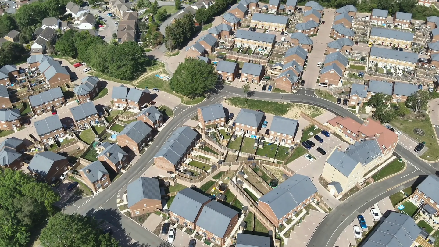 Aerial view of curved residential streets lined with modern houses, private gardens and small planted green spaces between homes.