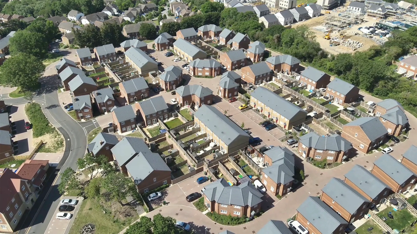 Oblique aerial view of a new housing scheme with brick homes, enclosed rear gardens and a construction site visible in the background.