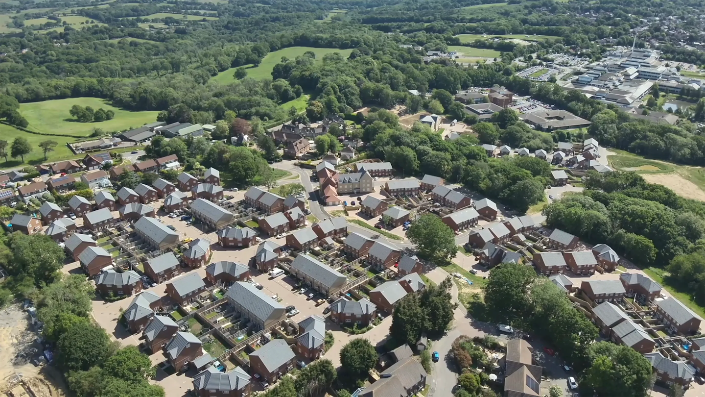 Aerial view of a completed housing development set within green countryside, showing tree-lined boundaries, surrounding fields and a nearby school campus.