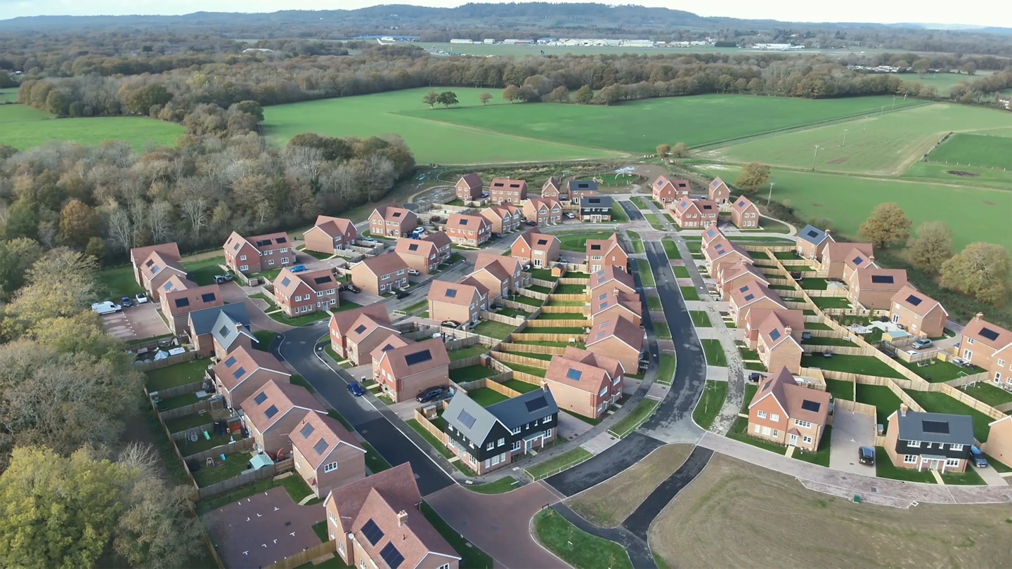 Aerial view of a completed housing development with detached and terraced homes, set among fields and woodland with curving roads.