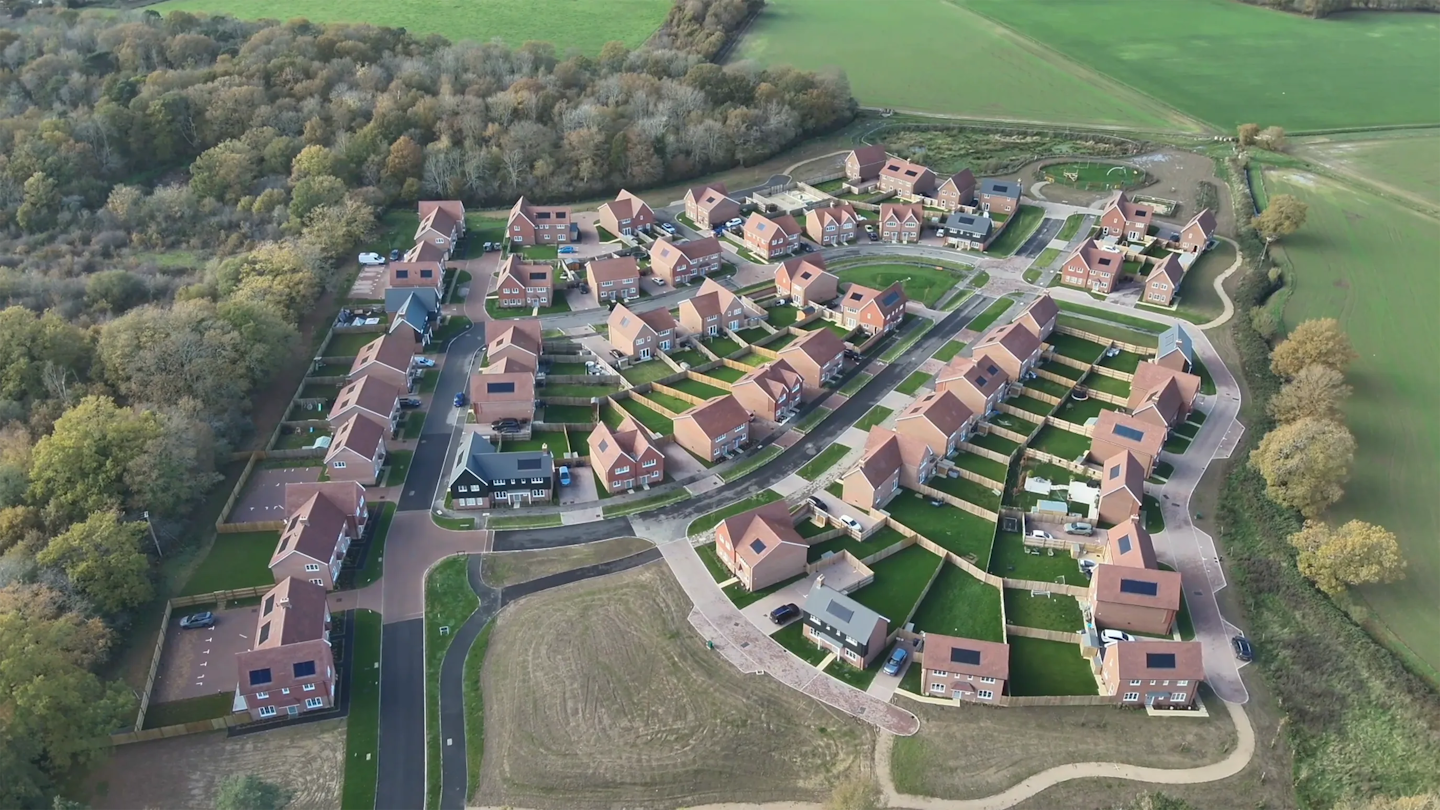 Wide aerial view of the development set within fields and woodland, with a clear street pattern and green space at its centre.