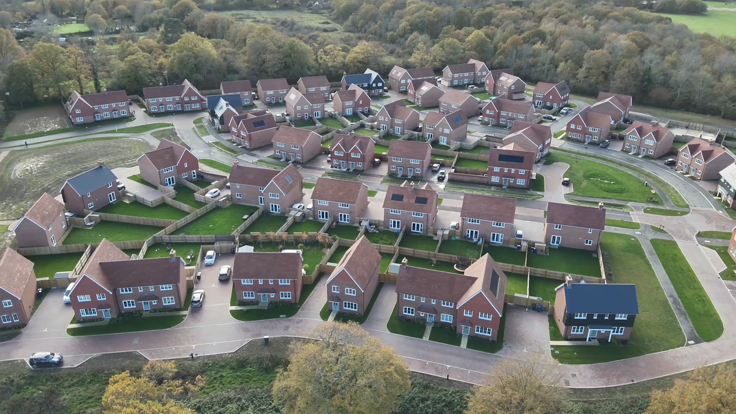Low aerial view showing a mix of detached and semi-detached homes with private gardens, driveways and planted verges.