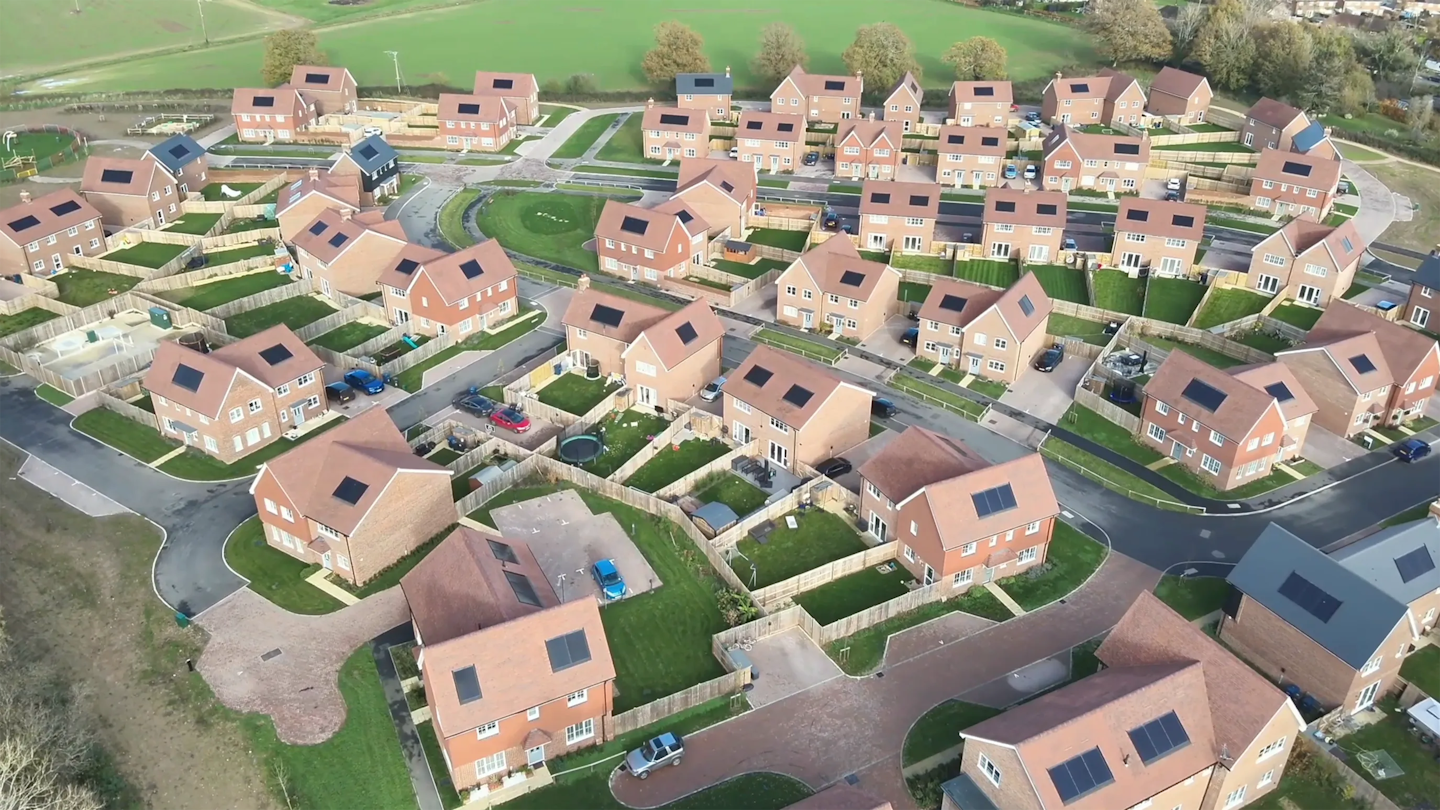 Aerial view of a residential area bordered by countryside, showing varied house types, rear gardens and a central road layout.