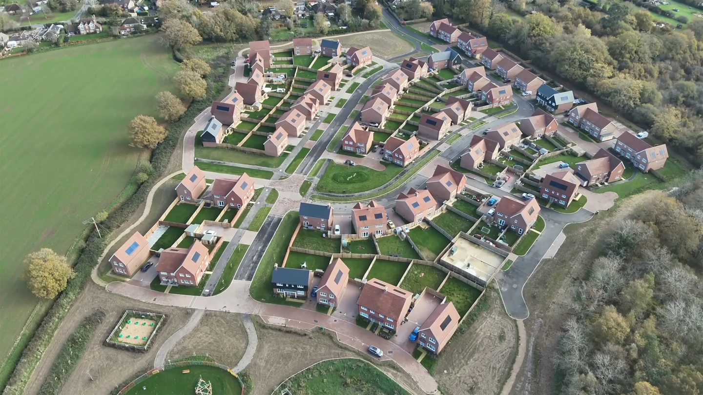Aerial view of a residential area bordered by countryside, showing varied house types, rear gardens and a central road layout.