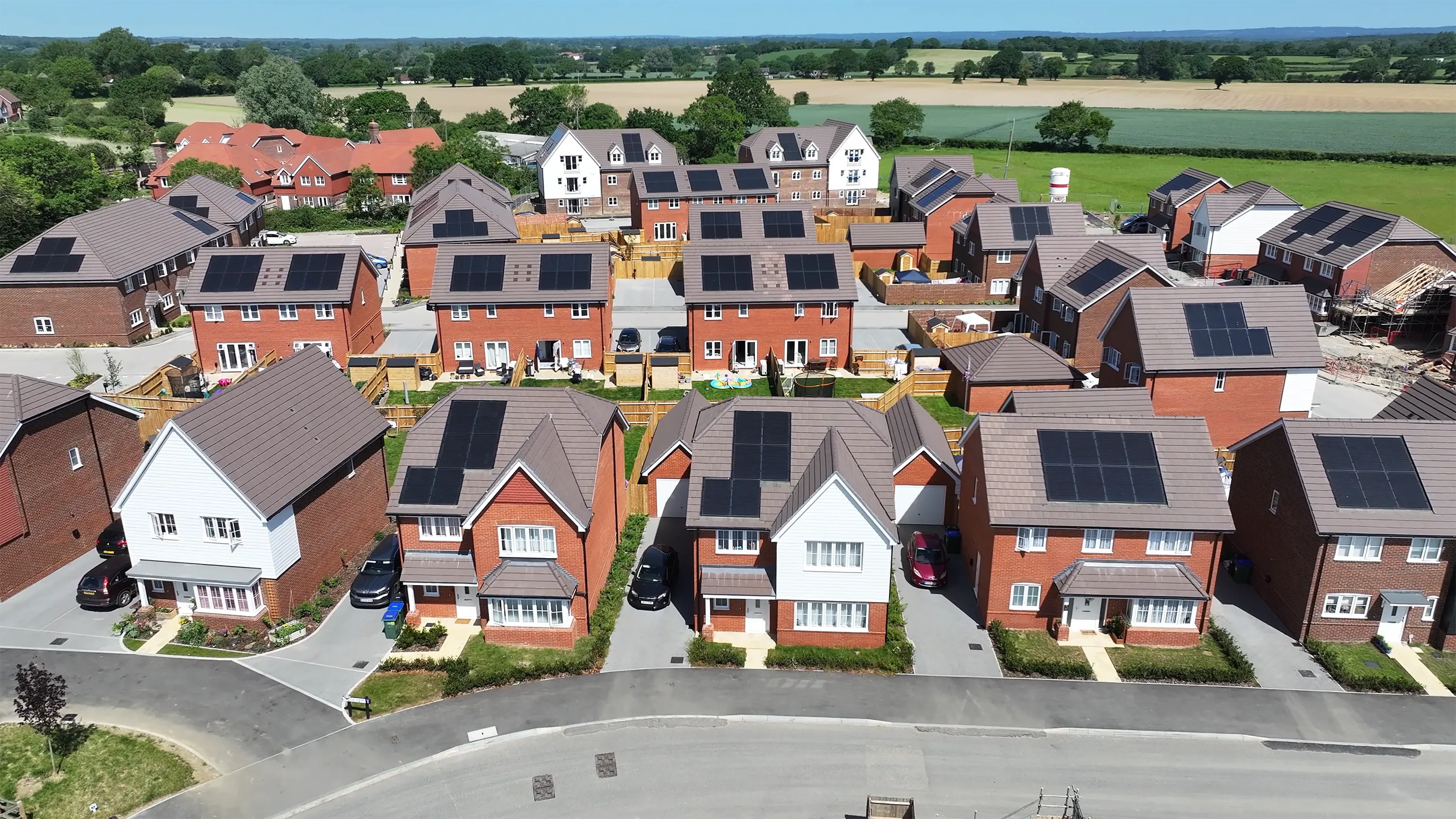 Aerial view showing a row of houses with solar panels, enclosed gardens and a backdrop of fields and existing village housing.