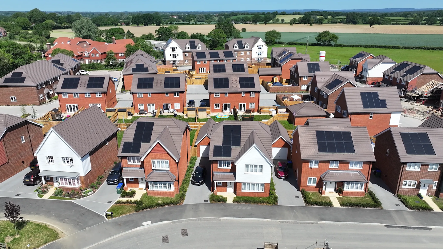 Aerial view showing a row of houses with solar panels, enclosed gardens and a backdrop of fields and existing village housing.
