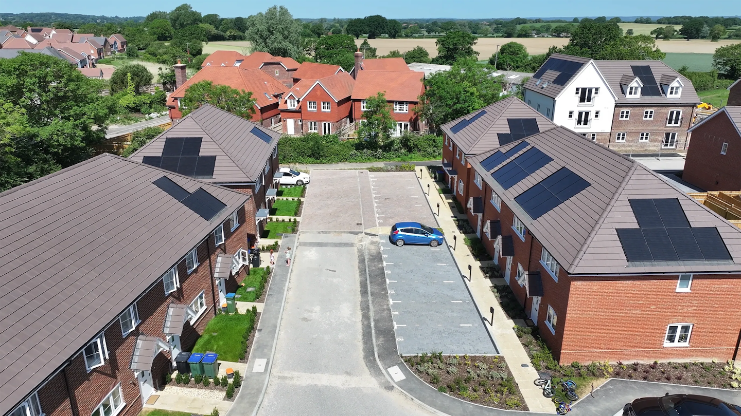 Street-level aerial view along a new road with brick homes on both sides, parking spaces and a few bicycles visible near the pavement.