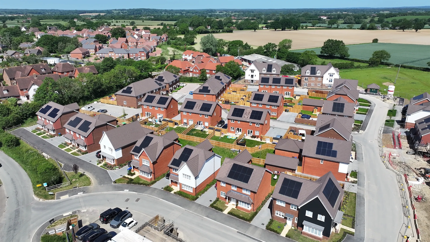 Aerial view of a completed housing development with semi-detached and detached homes, many with roof-mounted solar panels, set beside open countryside.