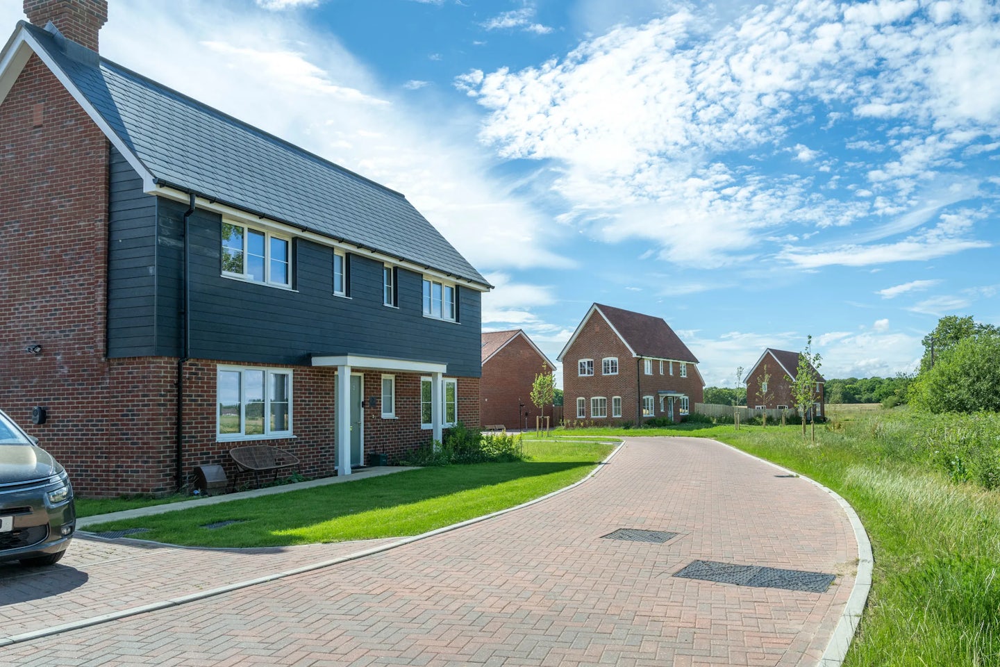 Detached and semi-detached red brick houses with driveways on a quiet road beside open fields.