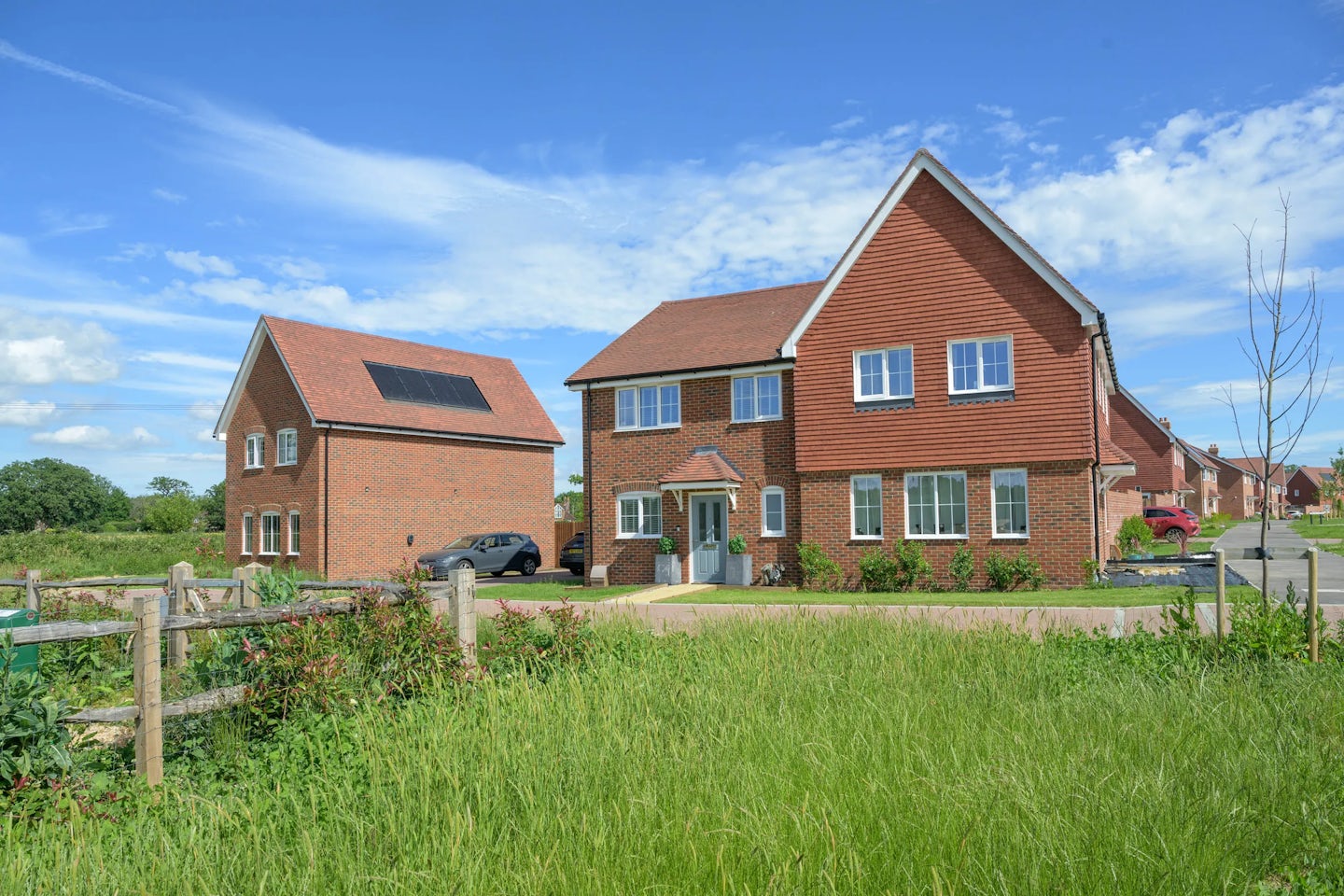 Detached red brick houses with driveways and lawns set beside open green space.
