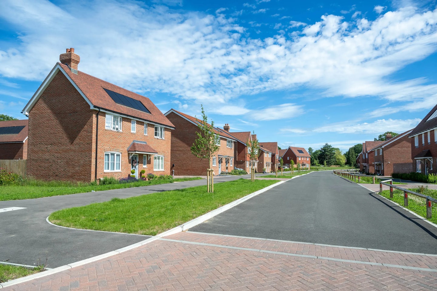 Street view of red brick houses with small front gardens, lawns and young trees along a wide road.