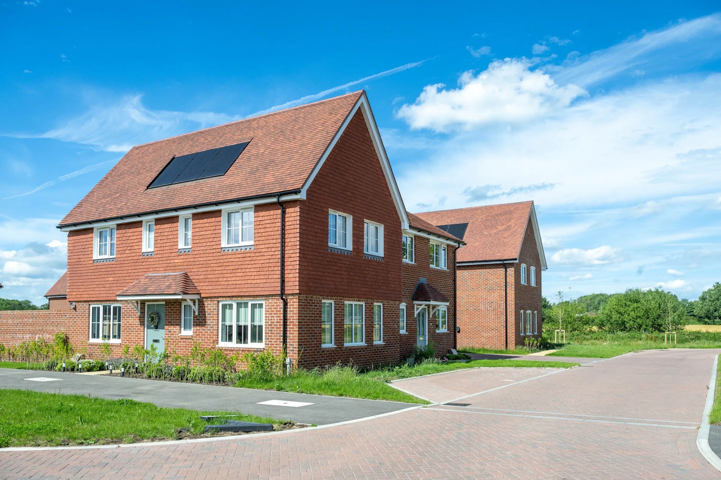 Pair of red brick houses with tiled roofs and solar panels, set on a corner with paved road and grass verge.