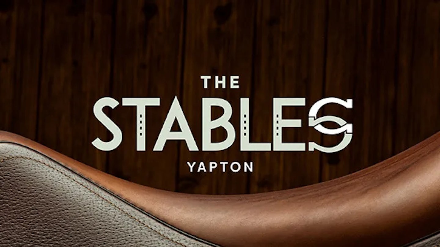 Modern logo for “The Stables Yapton” on a wood background, with a curved leather saddle detail in the foreground