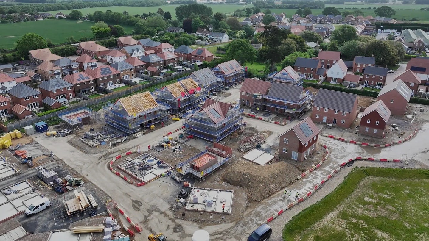 Construction site with multiple homes at different build stages, bordered by completed houses and greenery