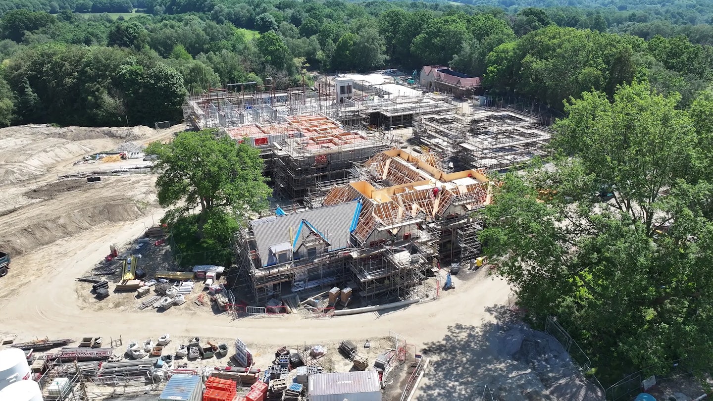 Aerial view of a construction site surrounded by trees, with multiple buildings under construction, scaffolding in place and materials stored around a curved access road.