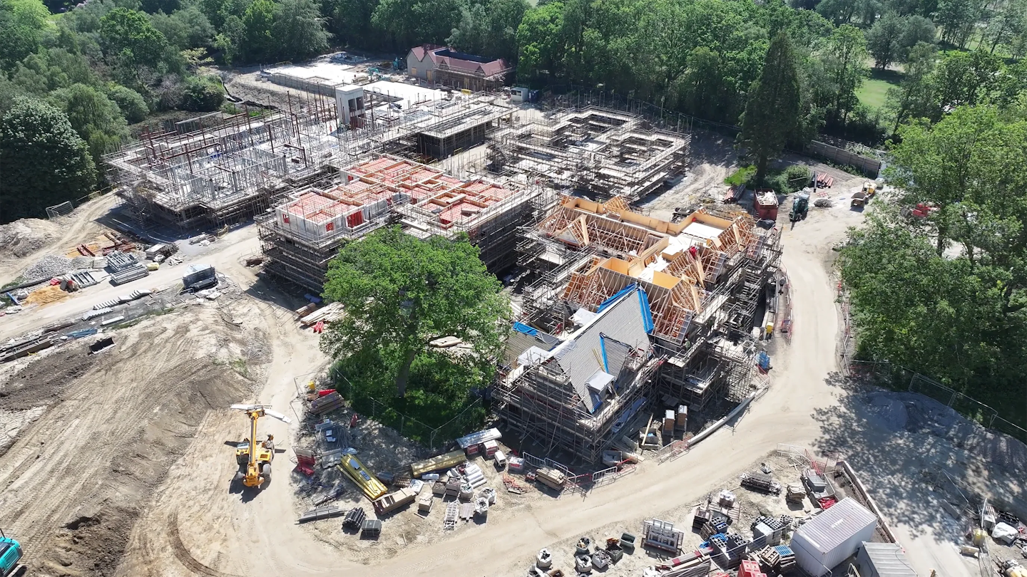 Aerial view of a multi-building construction site, with roof frames being installed on one block and other structures at earlier stages, set within a wooded landscape.