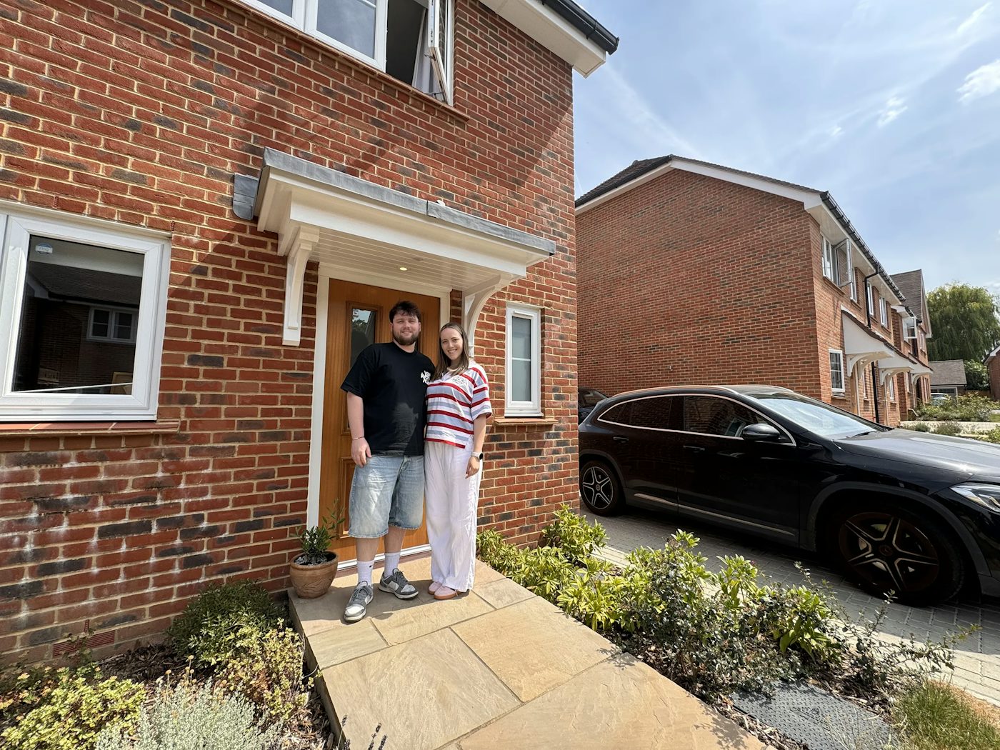 A couple stand in front a new build brick home. A black car is in the driveway.