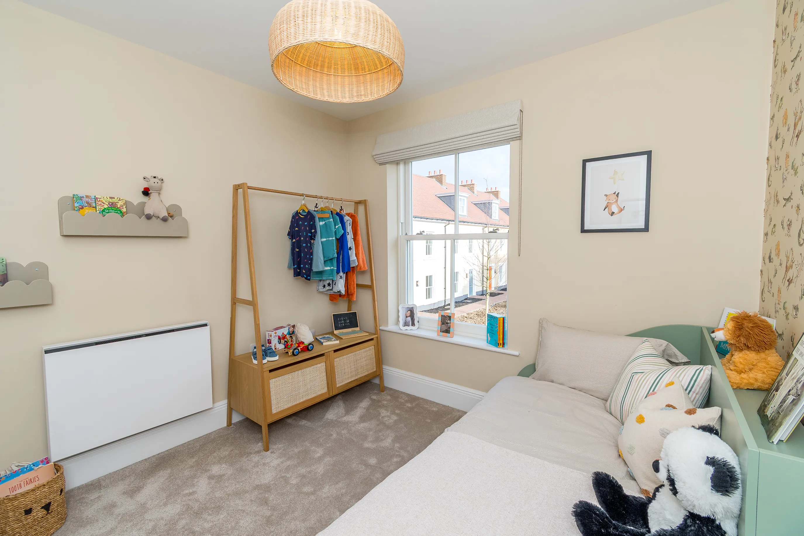 A childs bedroom with cream walls a white sash window and a low bed with plush animals and books.