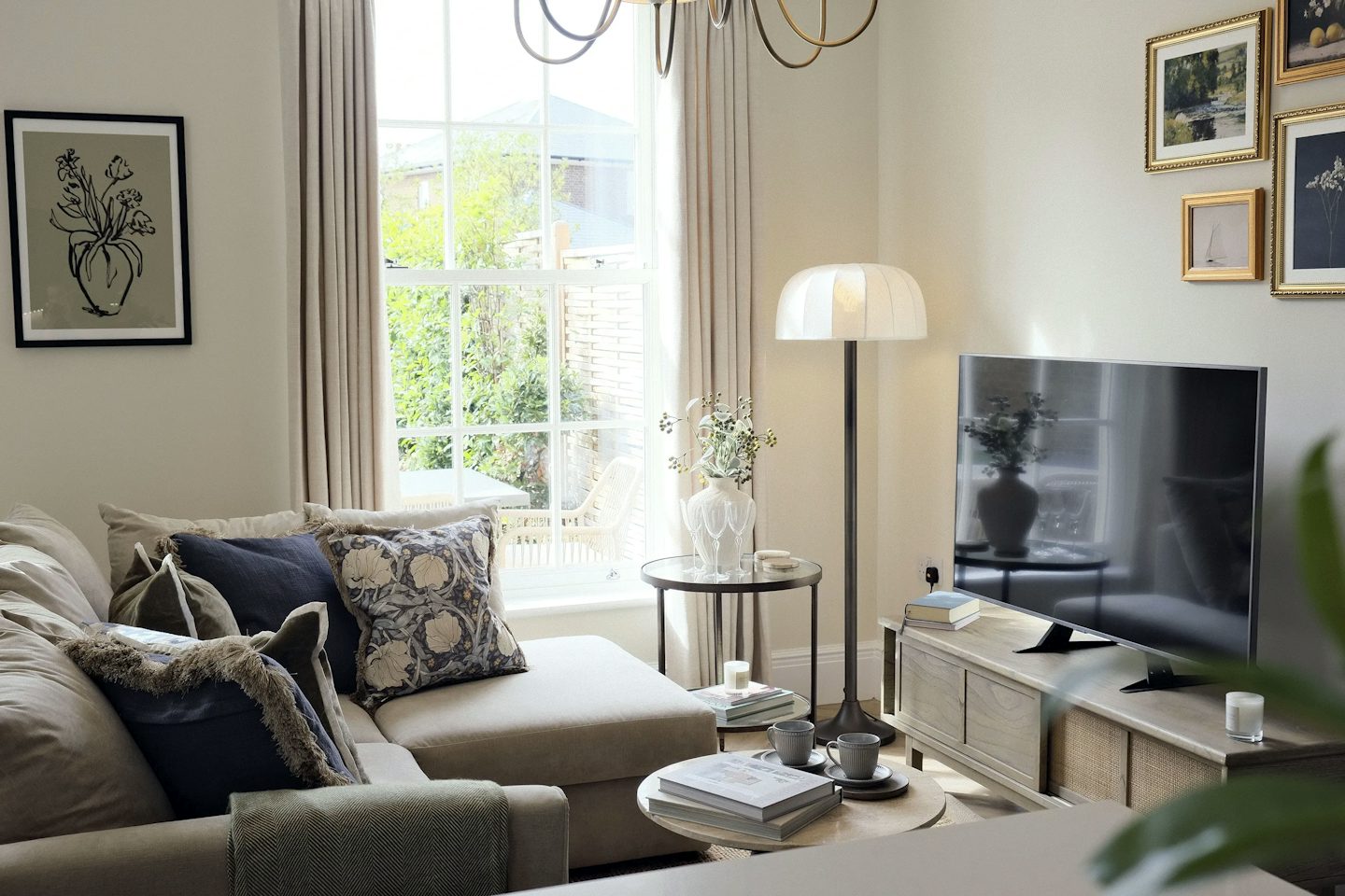 Comfortable living room featuring a large sofa, television, floor lamp, coffee table, and a window overlooking garden greenery.