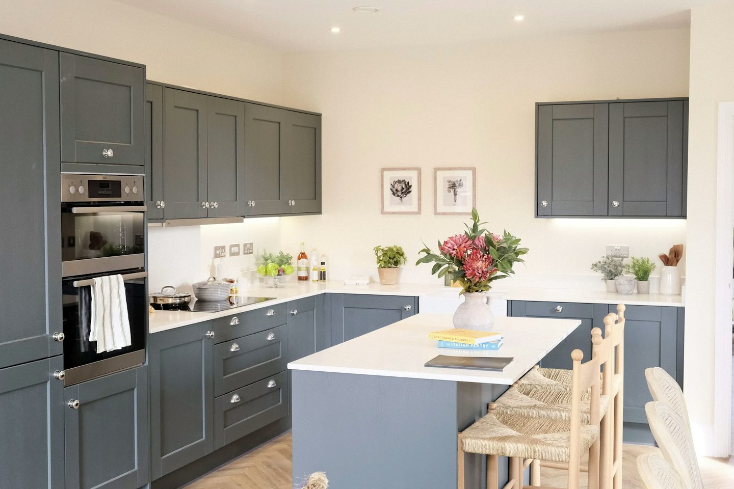 Contemporary kitchen with dark blue shaker-style cabinets, a central island with seating, white worktops, and decorative flowers on the island.