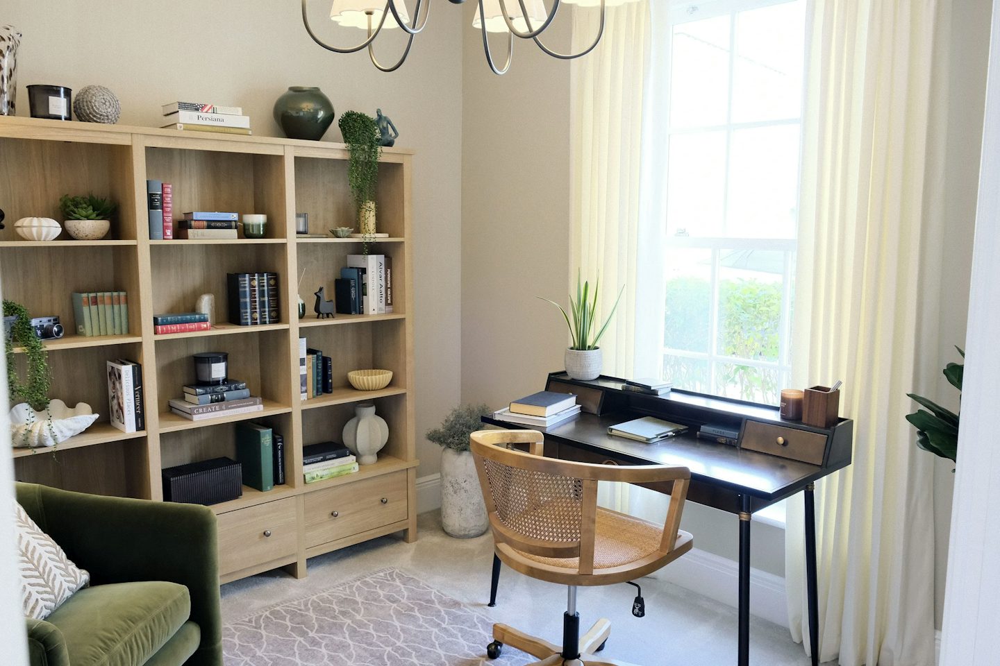 Home office with a wooden bookshelf filled with books and decorative items, a black desk, woven chair, and large window with sheer curtains.