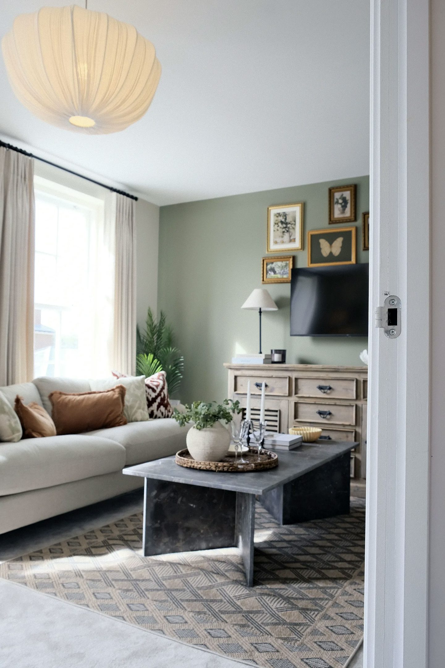 Bright living room with a cream sofa, marble coffee table, green feature wall, wall-mounted television, framed artwork, and large window with curtains.