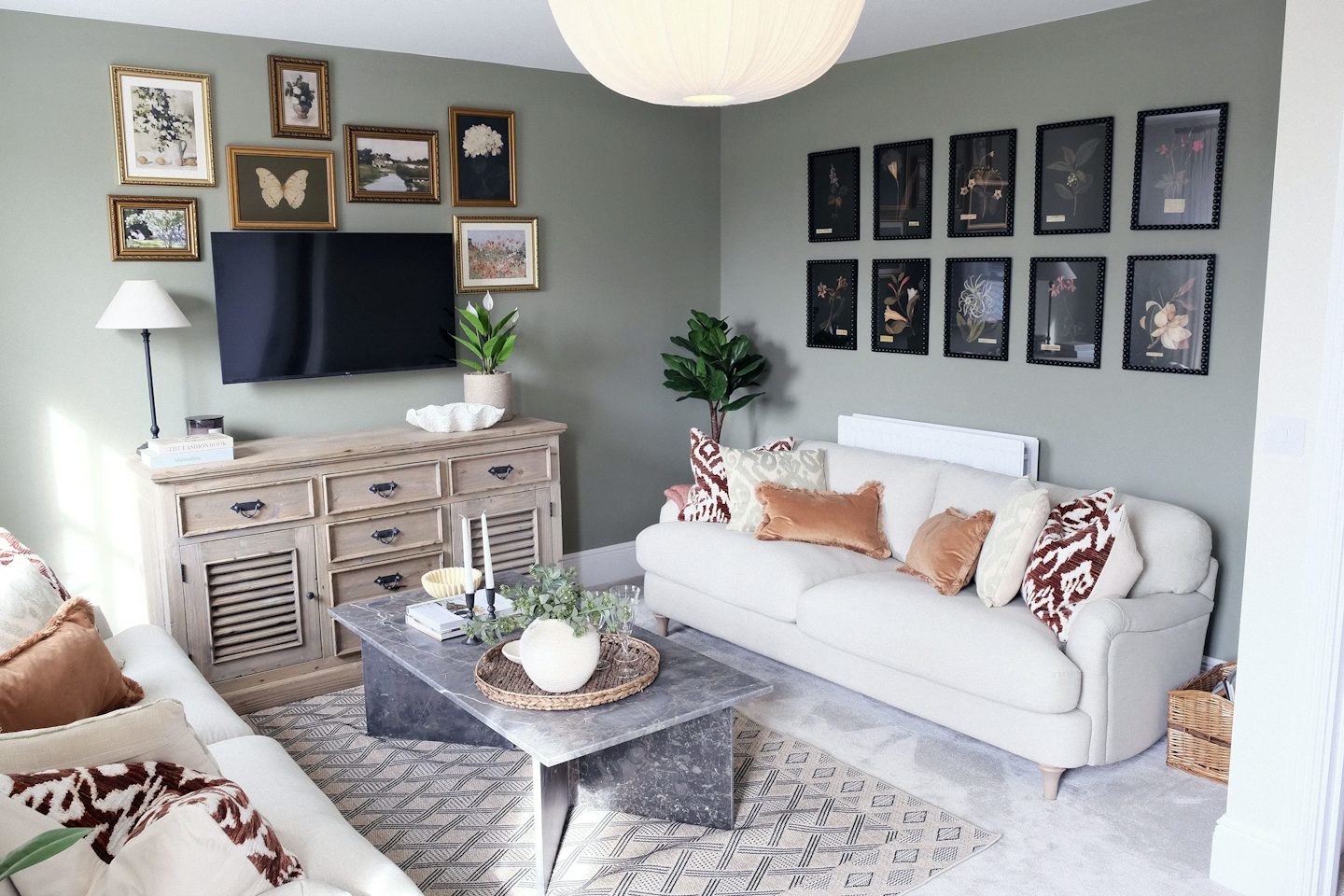 Contemporary living room with two cream sofas, a marble coffee table, botanical wall art, a television, and soft green walls.