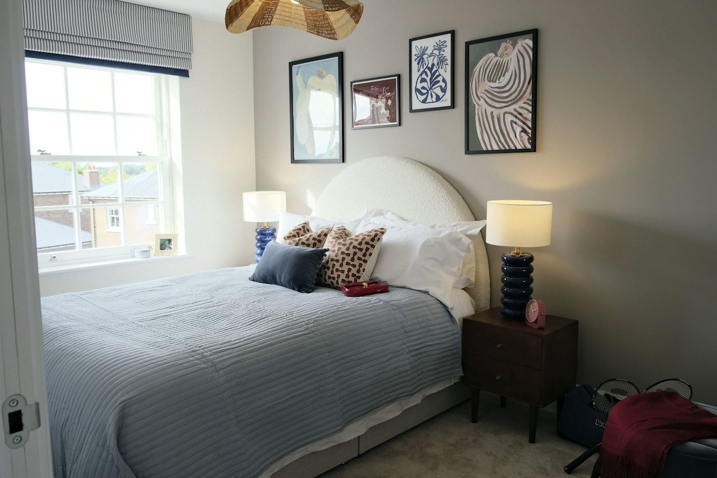 Modern double bedroom with a curved upholstered headboard, blue bedspread, bedside tables with lamps, framed artwork, and a large window providing natural light.
