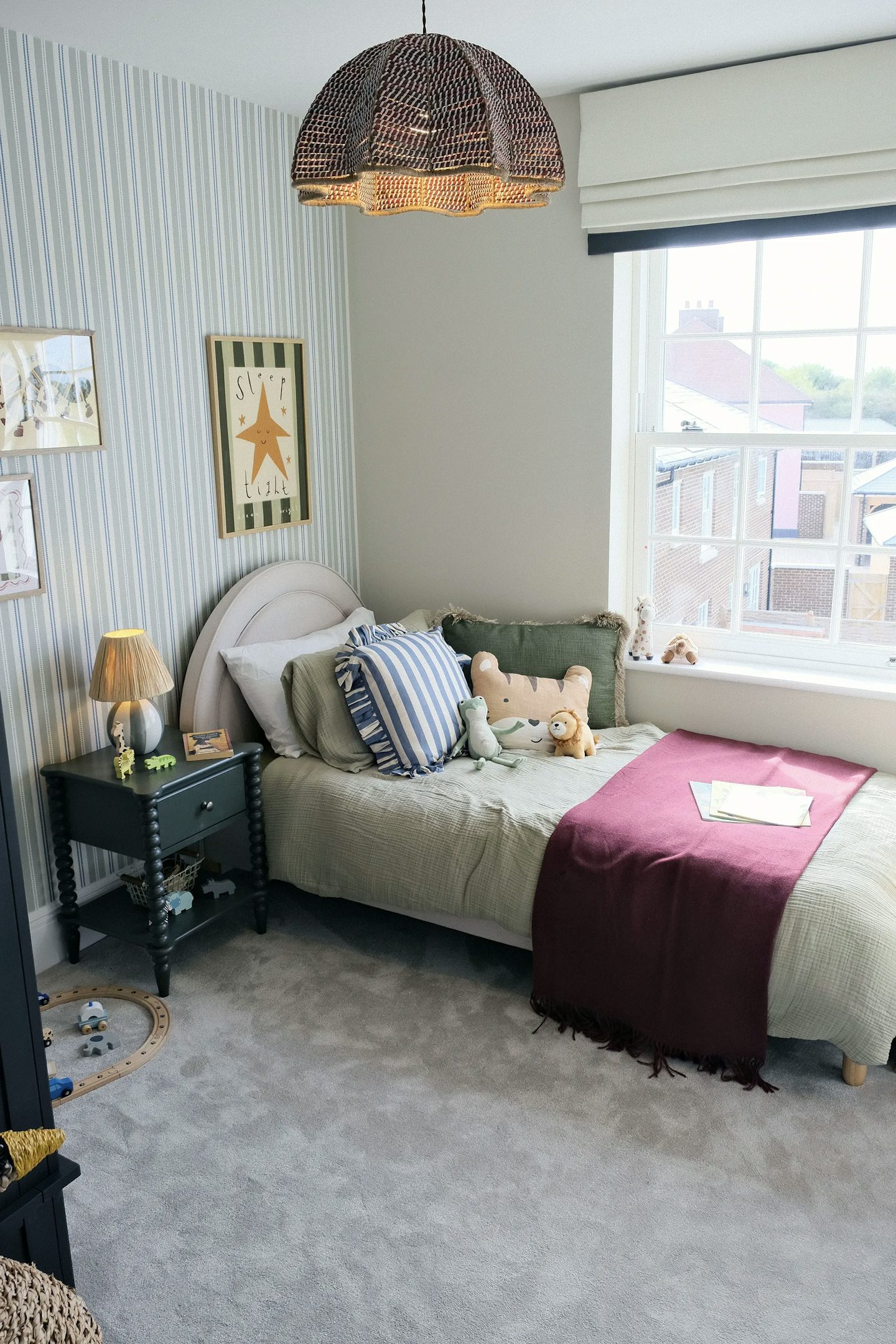 Children's bedroom with a single bed, striped feature wallpaper, colourful cushions and soft toys, bedside table with lamp, and a large window bringing in natural light.