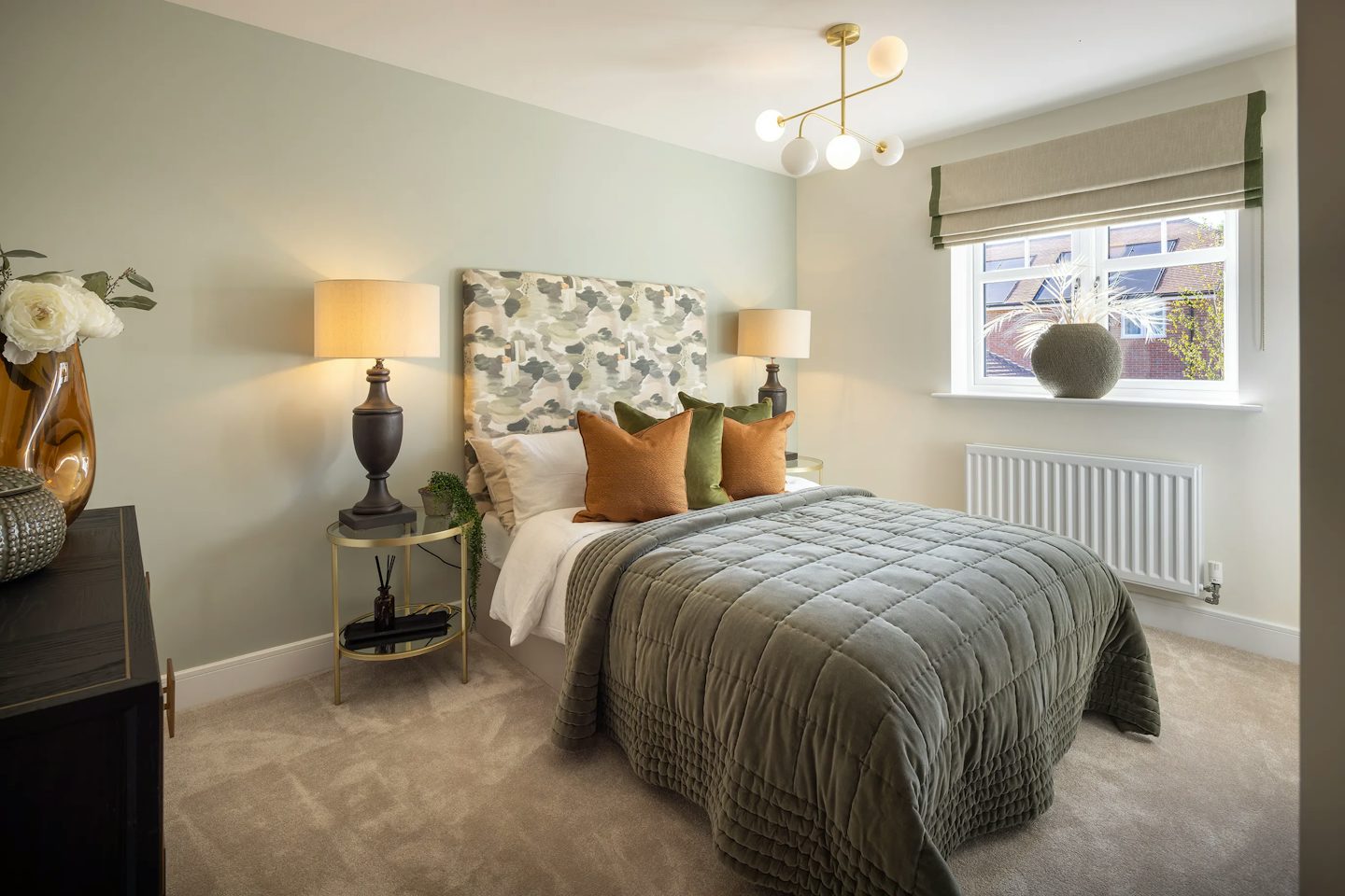 Contemporary bedroom with a patterned upholstered headboard, soft green walls, bedside tables with lamps, and a large window providing natural light.