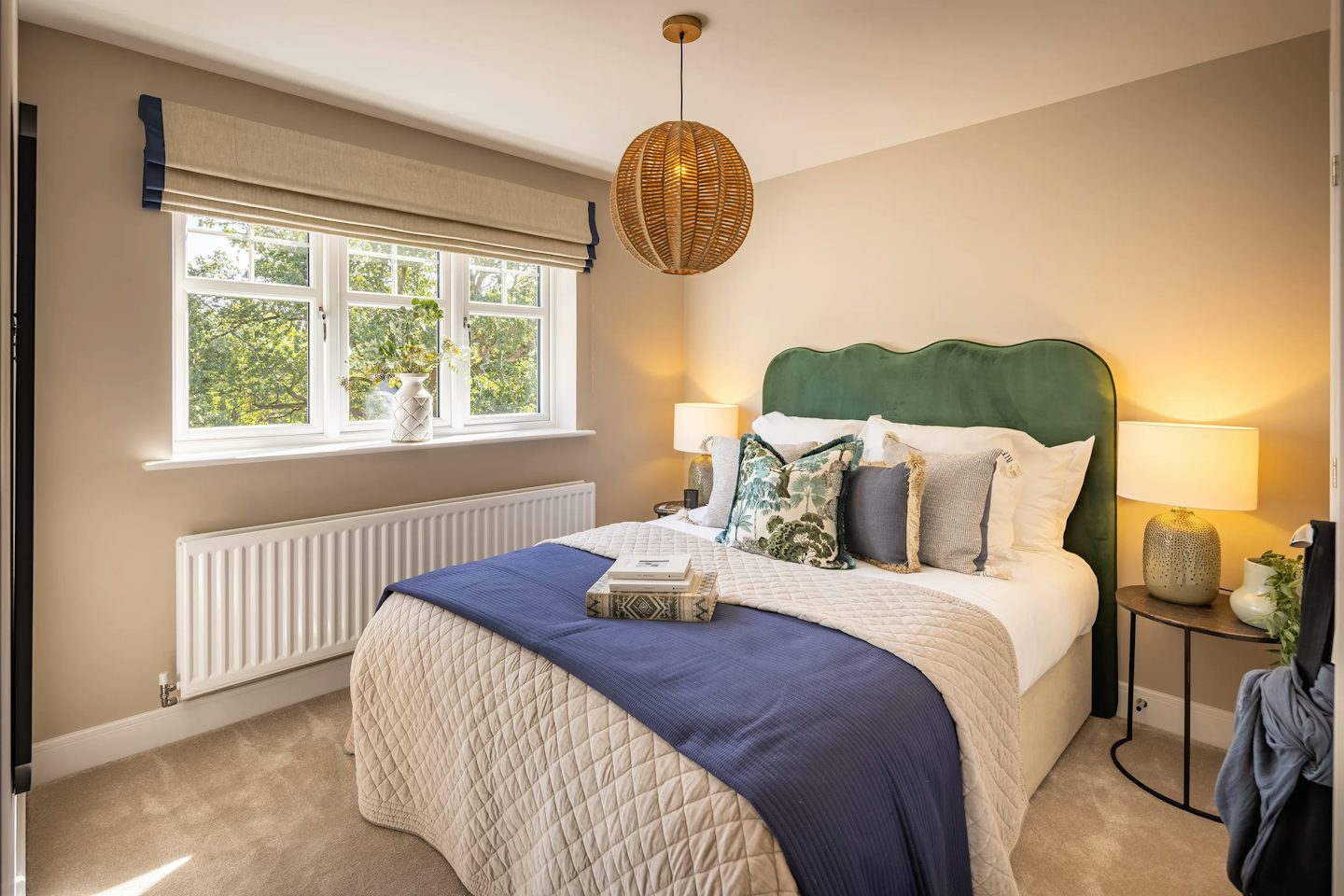 Stylish double bedroom with a green upholstered headboard, blue bed runner, bedside lamps, and a large window overlooking greenery.