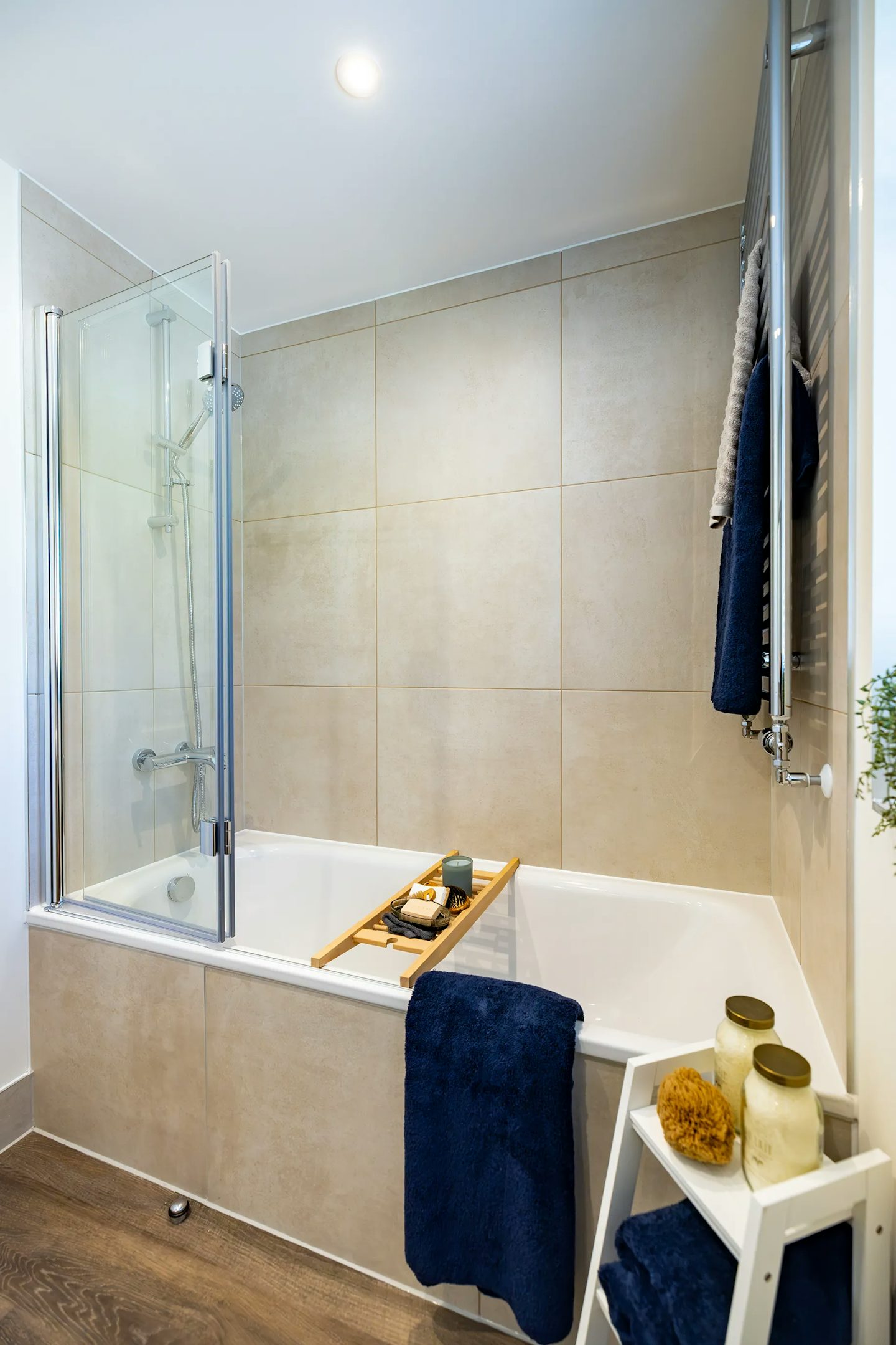 Modern family bathroom featuring a bath with glass shower screen, large neutral wall tiles, heated towel rail, and decorative accessories.