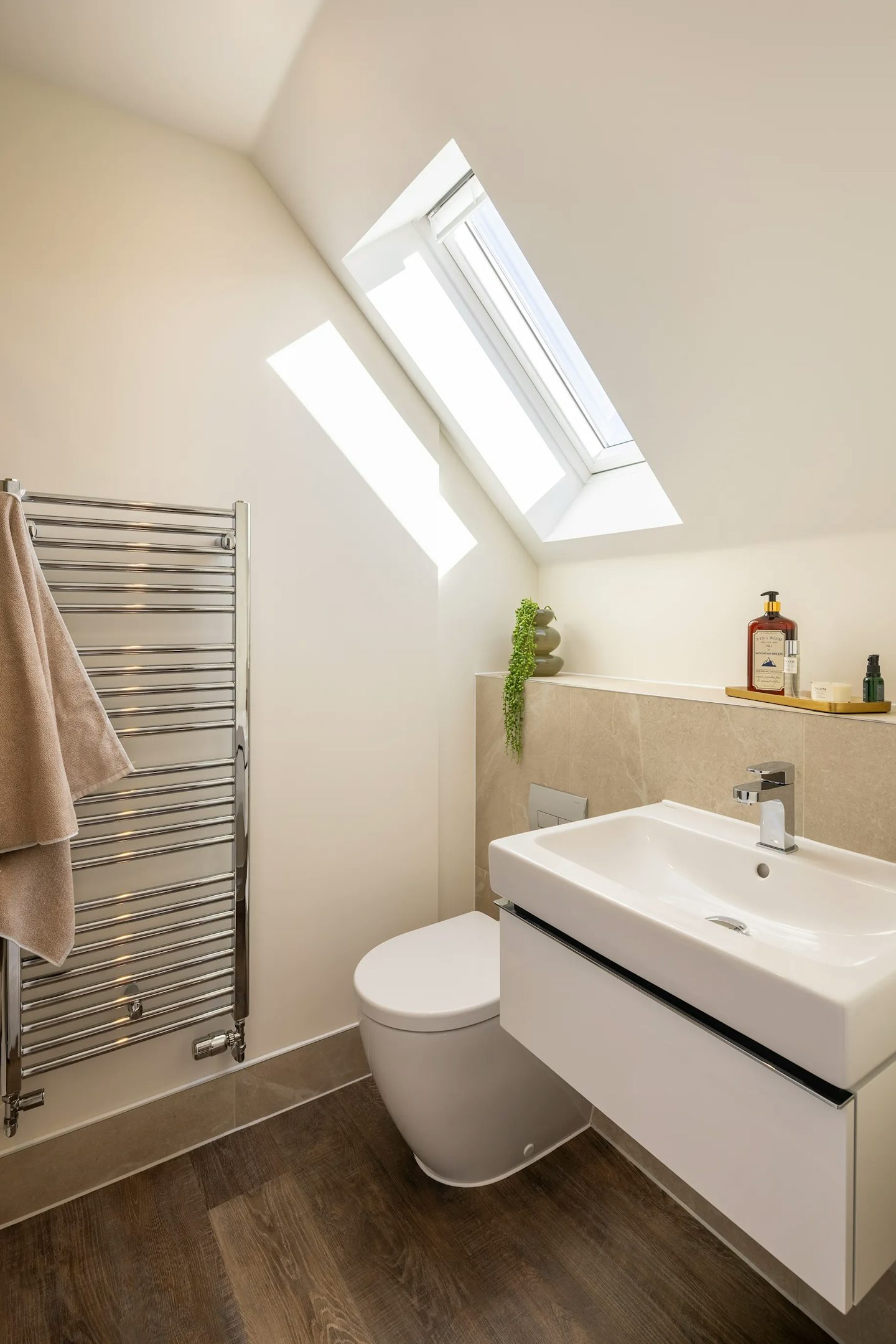 Contemporary bathroom with a skylight window, wall-mounted basin, toilet, chrome heated towel rail, and neutral tiled finishes.