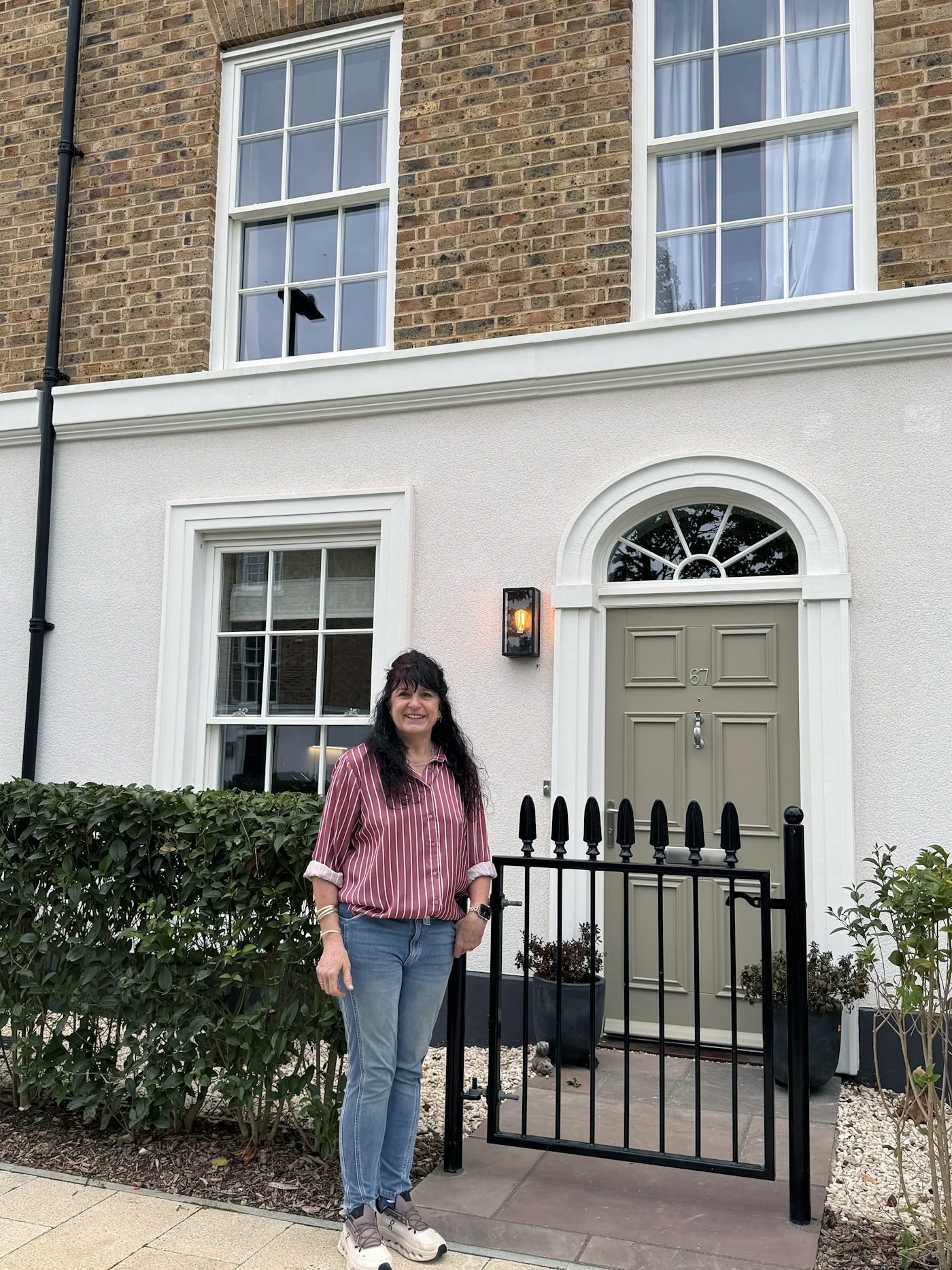 Person standing outside the entrance of a modern townhouse with brick and white-rendered exterior, landscaped frontage and black metal gate.