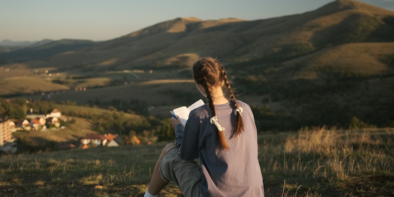 A girl reads a book overlooking a landscape.