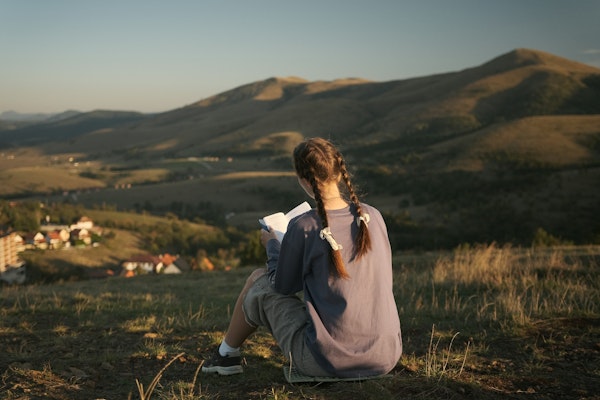 A girl reads a book overlooking a landscape.