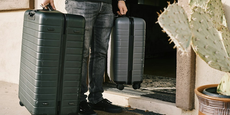 man in black denim jeans and black leather shoes standing beside black luggage bag