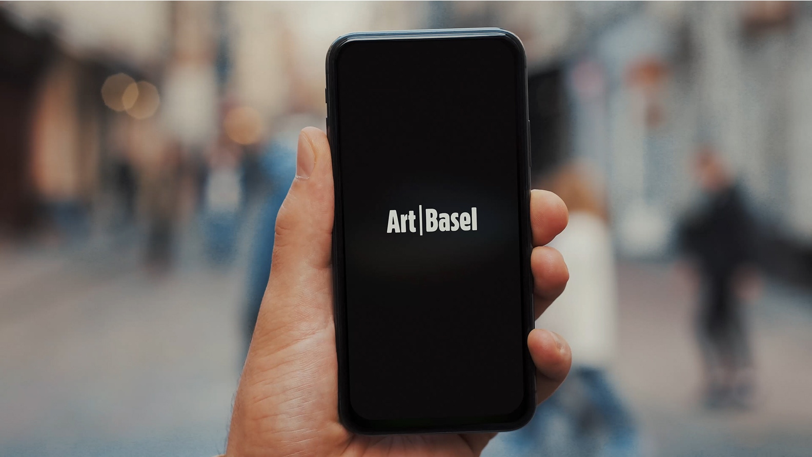 A hand holding a smartphone displaying the Art Basel logo on the screen, with a blurred city street background.