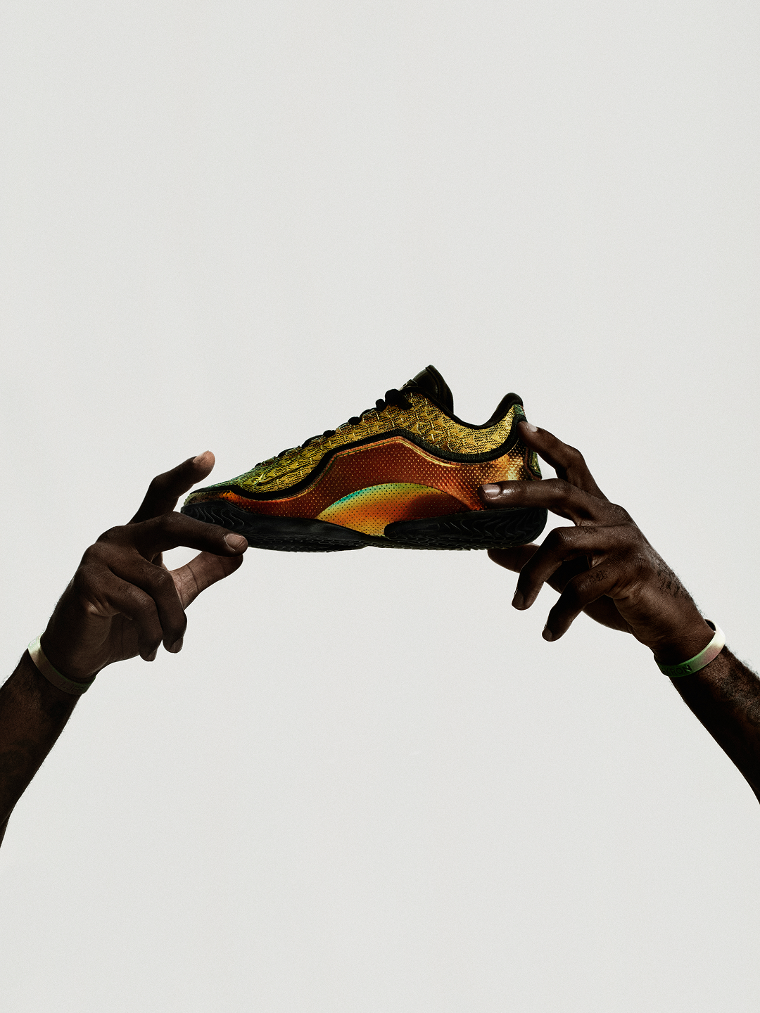 Two hands holding up a gold and orange iridescent LeBron XXIII shoe against a pale studio backdrop.