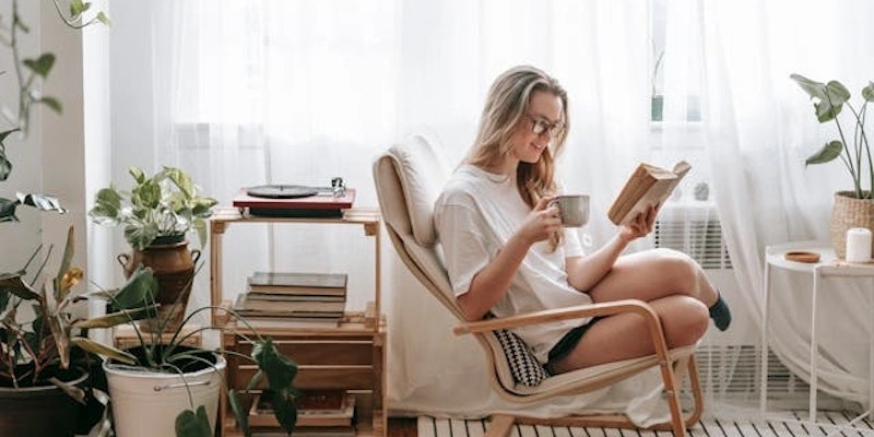 Imagem de uma mulher sorrindo, lendo um livro e tomando um café em frente a uma janela, evidenciando um bem-estar emocional.