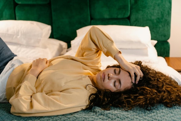 Mulher deitada na cama com a mão na testa e outra sobre o abdômen, representando sintomas como dor e mal-estar associados ao uso e indicações do Tropinal.