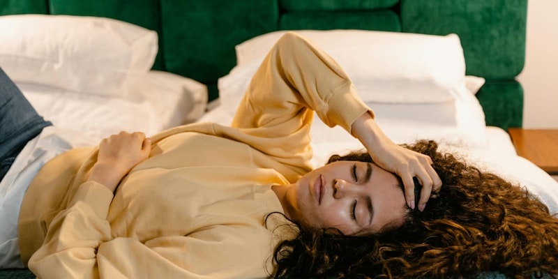Mulher deitada na cama com a mão na testa e outra sobre o abdômen, representando sintomas como dor e mal-estar associados ao uso e indicações do Tropinal.