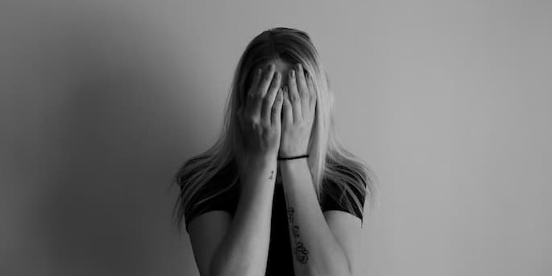 Mulher com as mãos cobrindo o rosto, em frente a uma parede lisa com ansiedade social tomando oxalato de escitalopram.