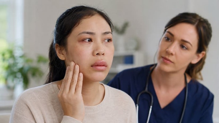 Paciente tocando o rosto com aparente inchaço na região dos lábios e da face durante consulta médica com angioedema alérgico.