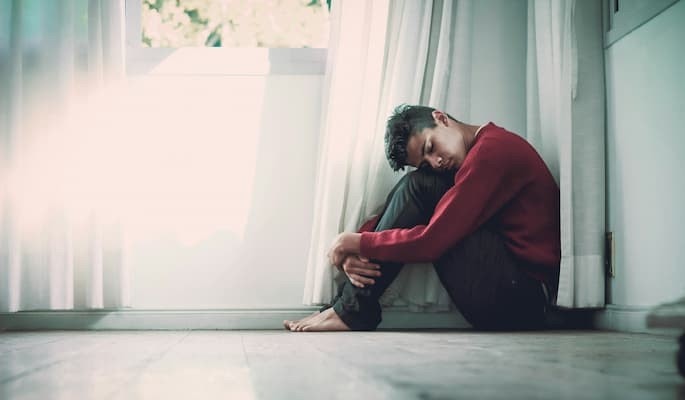 Pessoa sentada no chão, abraçando as pernas e com a cabeça apoiada nos joelhos, próxima a uma janela com transtorno depressivo maior (TDM).