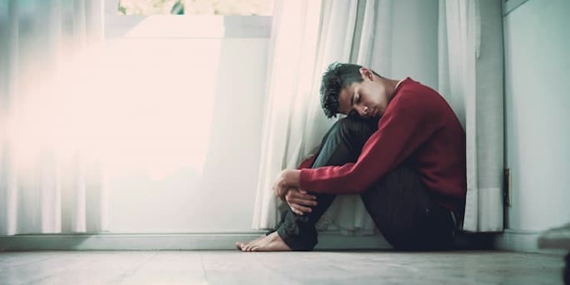 Pessoa sentada no chão, abraçando as pernas e com a cabeça apoiada nos joelhos, próxima a uma janela com transtorno depressivo maior (TDM).